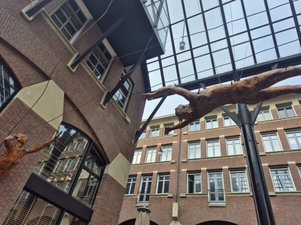 Onder de glazen overkapping bij Het Goudkantoor in Groningen hangen twee figuren. Sculptuur uit 1996, Farsi Largo/Making Space, is van kunstenaar Janet Mullarney